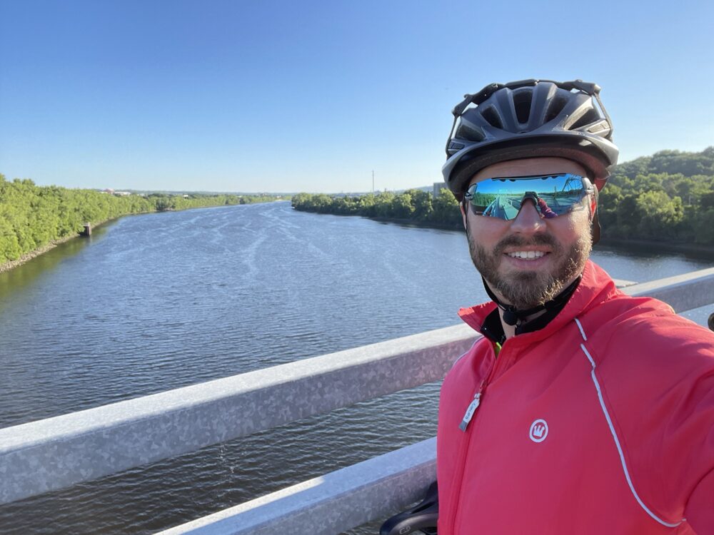 A man in a bicycle helmet, blue reflective sunglasses and a red windbreaker, in front of a railing overlooking a wide river, blueish gray in color with green banks.