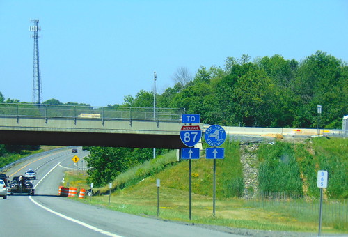 Highway with blue New York Thruway signs off to the right. Green grass and blue sky in the background.