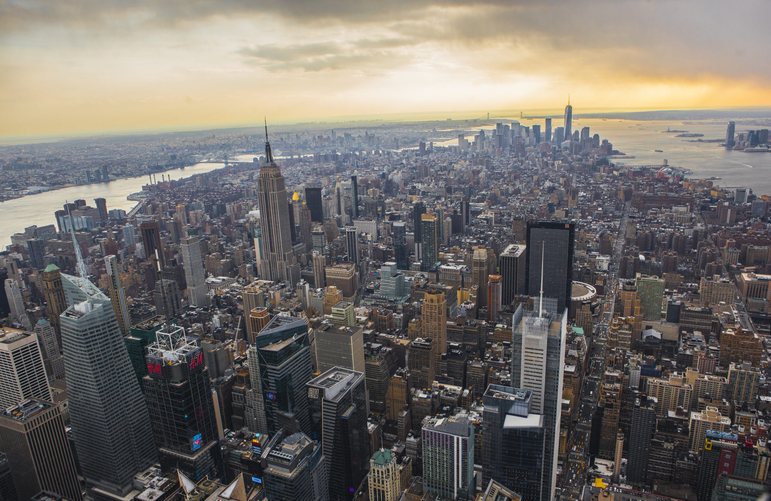 A view of Manhattan from high up, the buildings spreading out into the distance under a cloudy orange sky.