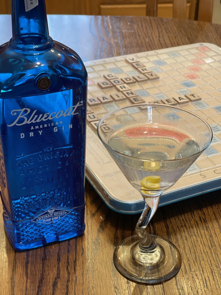 A clear martini in a martini glass, framed by a bright blue bottle of gin on its left, and an old, dusty, plastic Scrabble board with a few words on it on its right. 