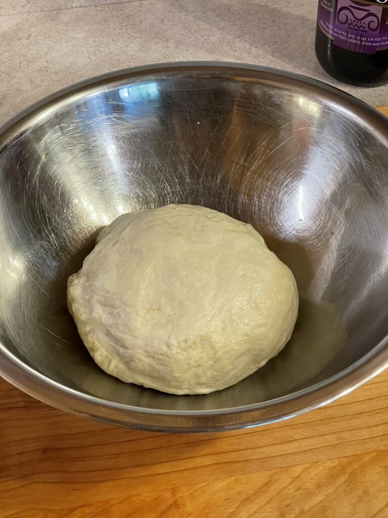 A silver mixing bowl on a wooden surface. It has a beige-colored, mostly round, ball of dough at the bottom.