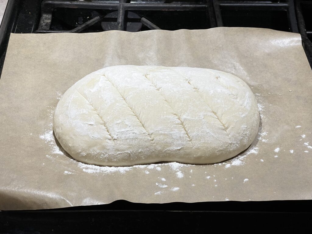 A loaf of bread on parchment paper, ready to go in the oven. It's covered in a dusting of white flour, and diagonal slashes cross the top of the loaf at regular intervals.