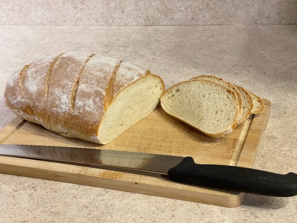 A loaf of bread sits on a wooden cutting board. It's partially sliced, with the slices stacked neatly on the right, leaving the remainder of the loaf on the left. A silver bread knife with a black handle rests on the cutting board in front of the bread.