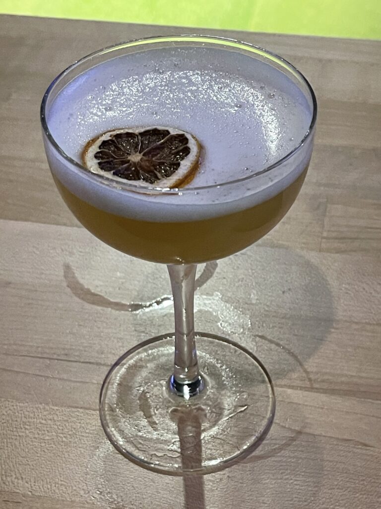 A coupe-style cocktail glass sits on a wooden surface. It's filled about halfway with an opaque yellowish liquid. A layer of white foam rests on top, and in the foam floats a circle of candied blood orange: nearly white pith forming a wagon-wheel pattern against the deep red of the flesh of the orange.