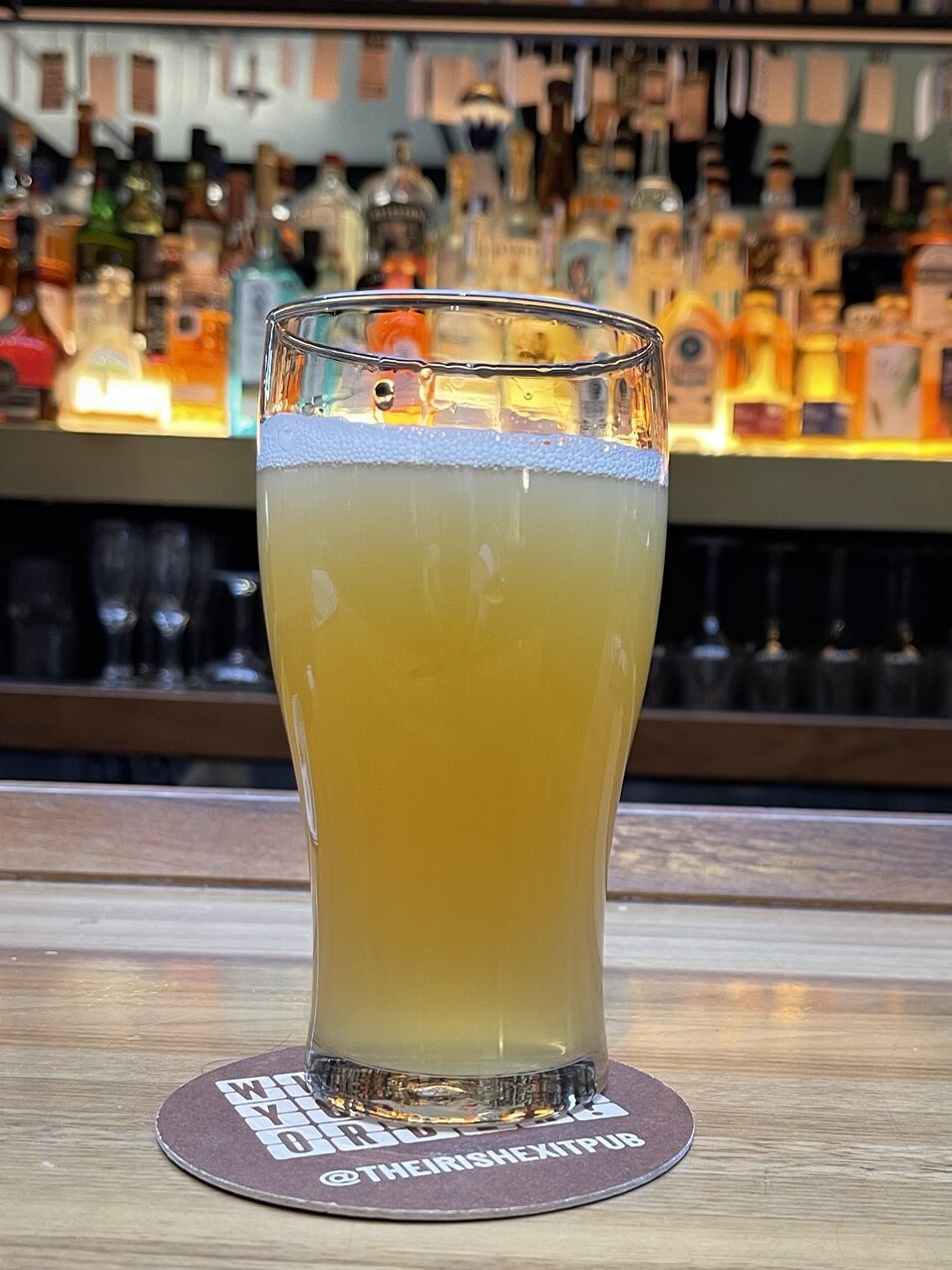 A pint glass sits on a bar. It's mostly full of an opaque pale golden liquid, with a layer of white bubbles on top. It's backlit by a shelf filled by many rows of bottles, lit from beneath with an orange-yellow light.