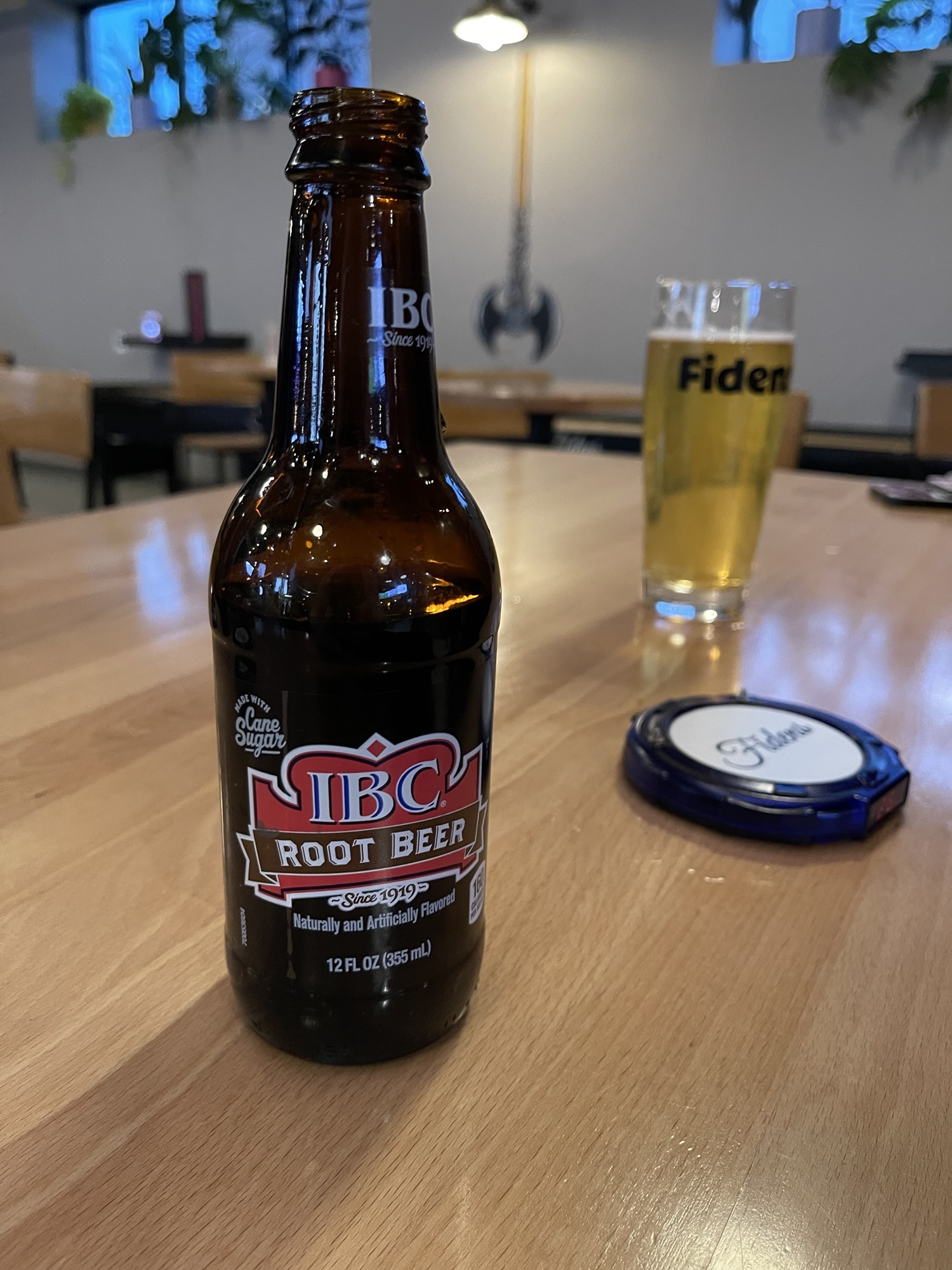A brown glass bottle and a beer glass sit on a wooden table. The bottle is in the foreground, and a red label with white lettering indicates IBC Root Beer. The beer glass is in the background, slightly out of focus. It's mostly full of a pale golden-colored beer with a thin layer of foam on top. Black lettering on the glass spells out Fiden's.