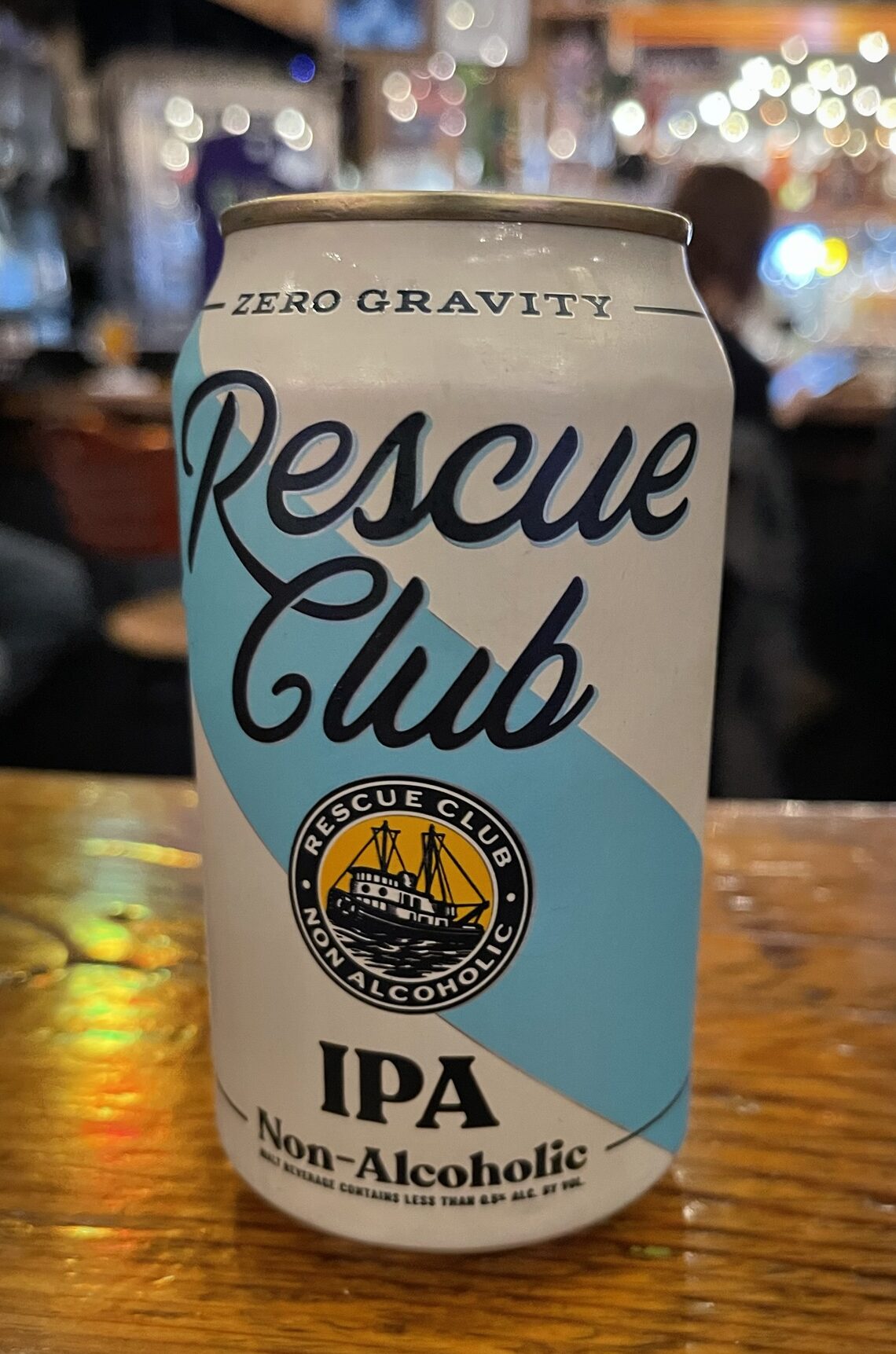 A close-up of a beer can siting on a wooden surface. It's white with a diagonal blue stripe and dark cursive lettering spelling out Rescue Club. In the background, out of focus are some people sitting at a bar. Light reflecting off of bottles etc. creates a sparkling effect.