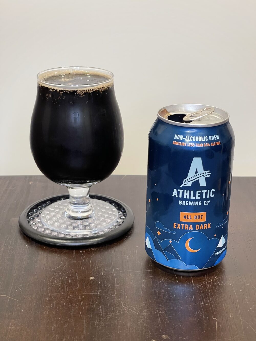 A tulip-shaped beer glass on a coaster and a beer can sit on a wooden table in front of an off-white background. The beer glass is full of a deep brown-colored liquid, almost black. A few bubbles are visible near the top around the rim of the glass. The beer can is blue, with a blue "A" shaped logo and the text ATHLETIC BREWING CO. Yellow text beneath indicate that the can contains ALL OUT Extra Dark.