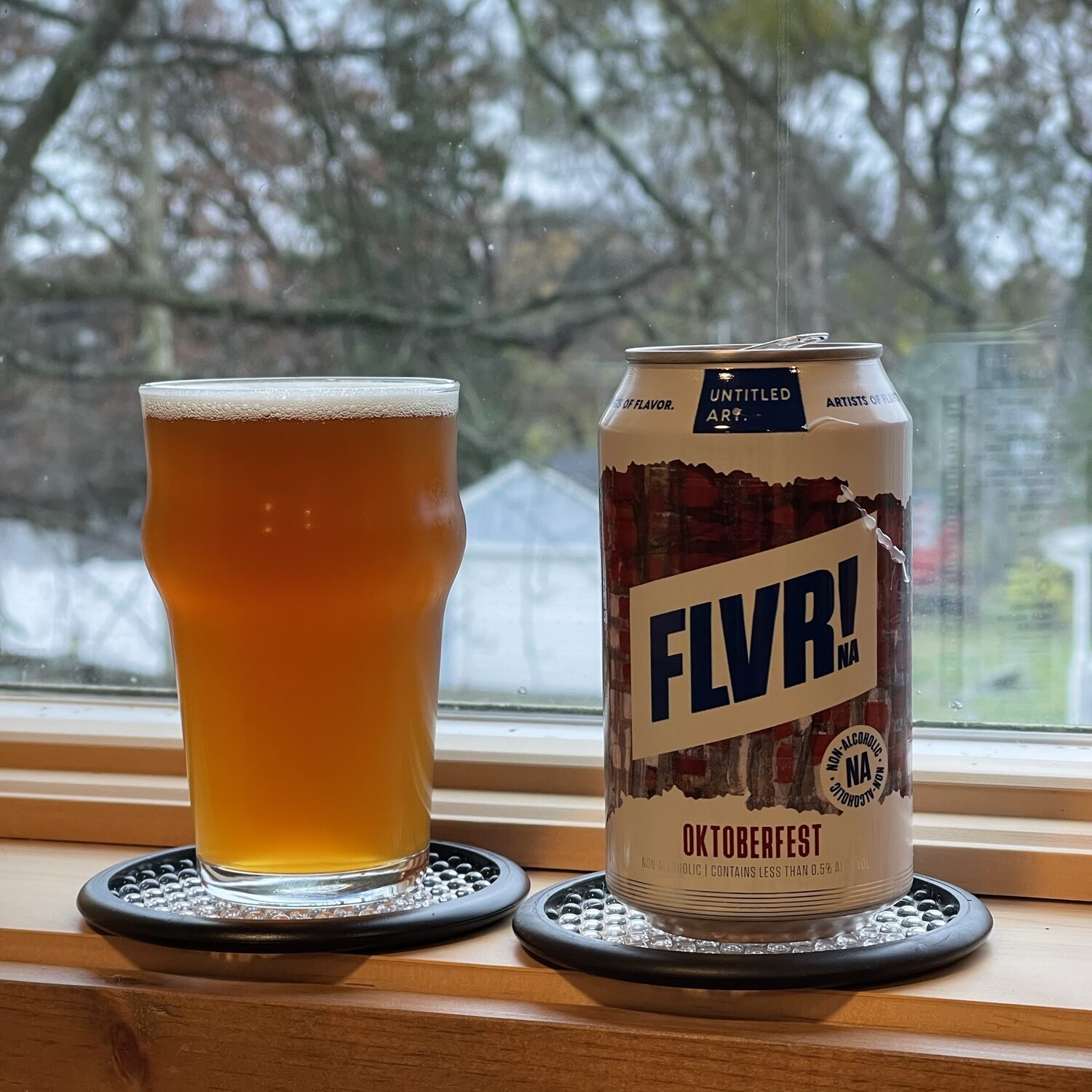 A beer can and a small beer glass sit on a windowsill. The glass is full of a deep amber colored liquid, a thin layer of white foam on top. The can is white with a reddish pattern across its middle. Blue and red text indicate that it's FLVR! Oktoberfest.