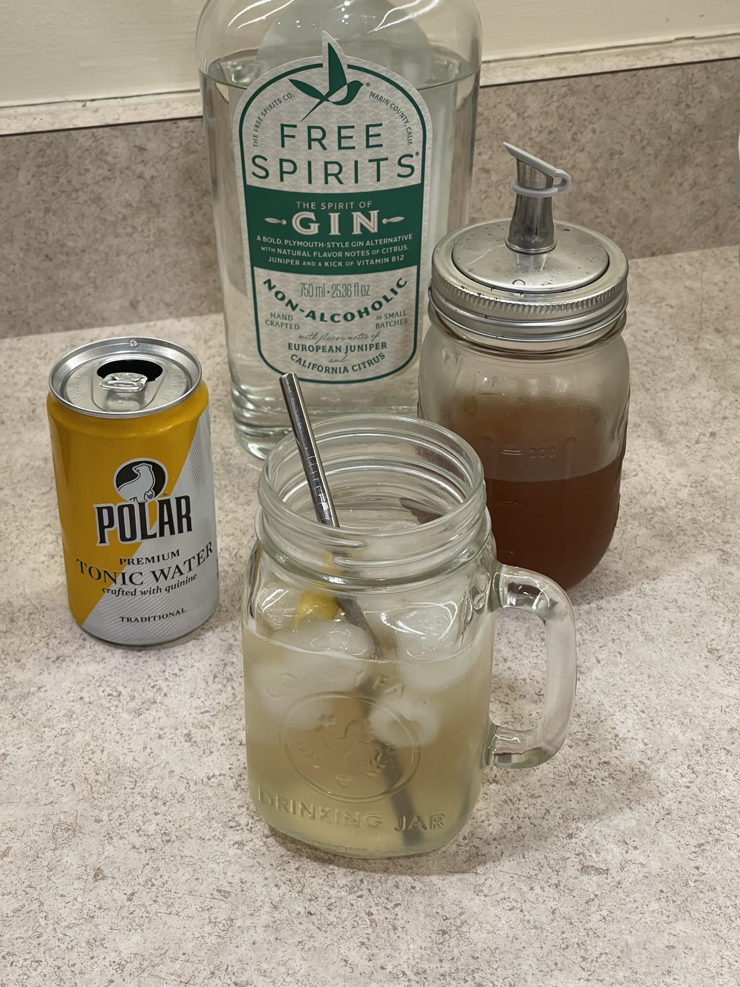 A mason jar with an iced beverage in it sits on a countertop in front of a liquor bottle, a small aluminum can, and a jar with a golden colored syrup.