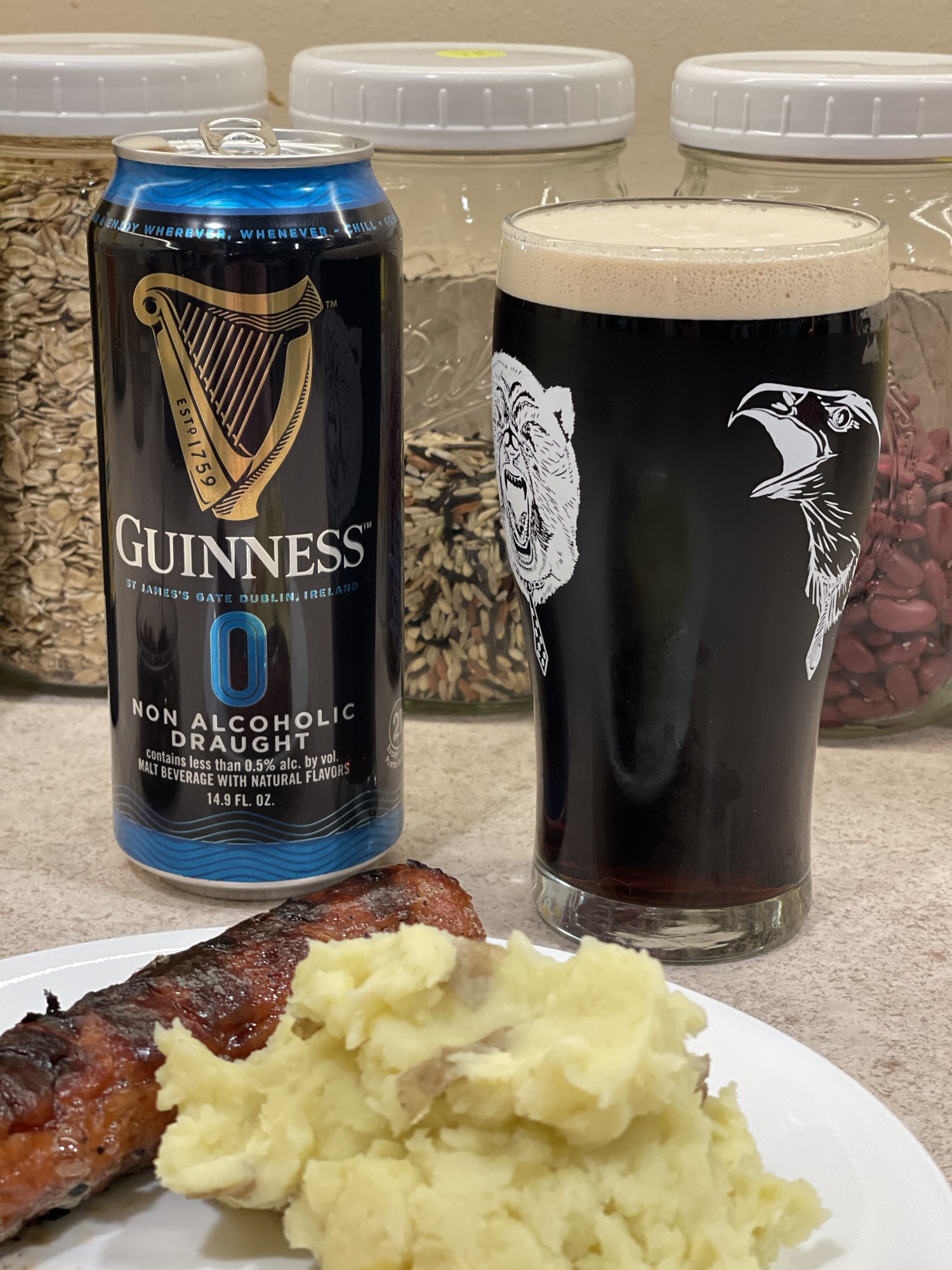 A beer can and full pint glass on a kitchen counter. Some food appears in the foreground, and some jars in the background. The beer can is black with some blue trim. The harp logo of the Guinness brand shows on the front with Guinness spelled out beneath. The pint glass is full of a deep black liquid, with a layer of light colored foam on top.