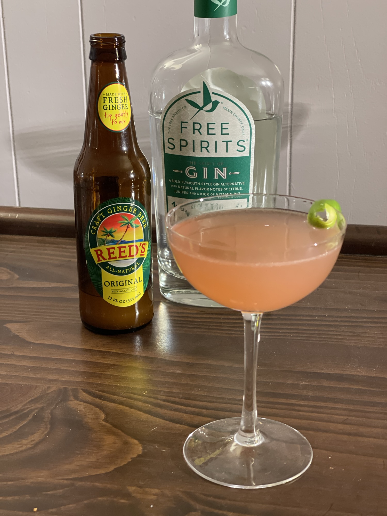 A coupe-style cocktail glass sits on a wooden bar, with two bottles in the background. It's mostly full of a light orangish pink liquid, slightly translucent. A green twist of lime peel rests on its rim.