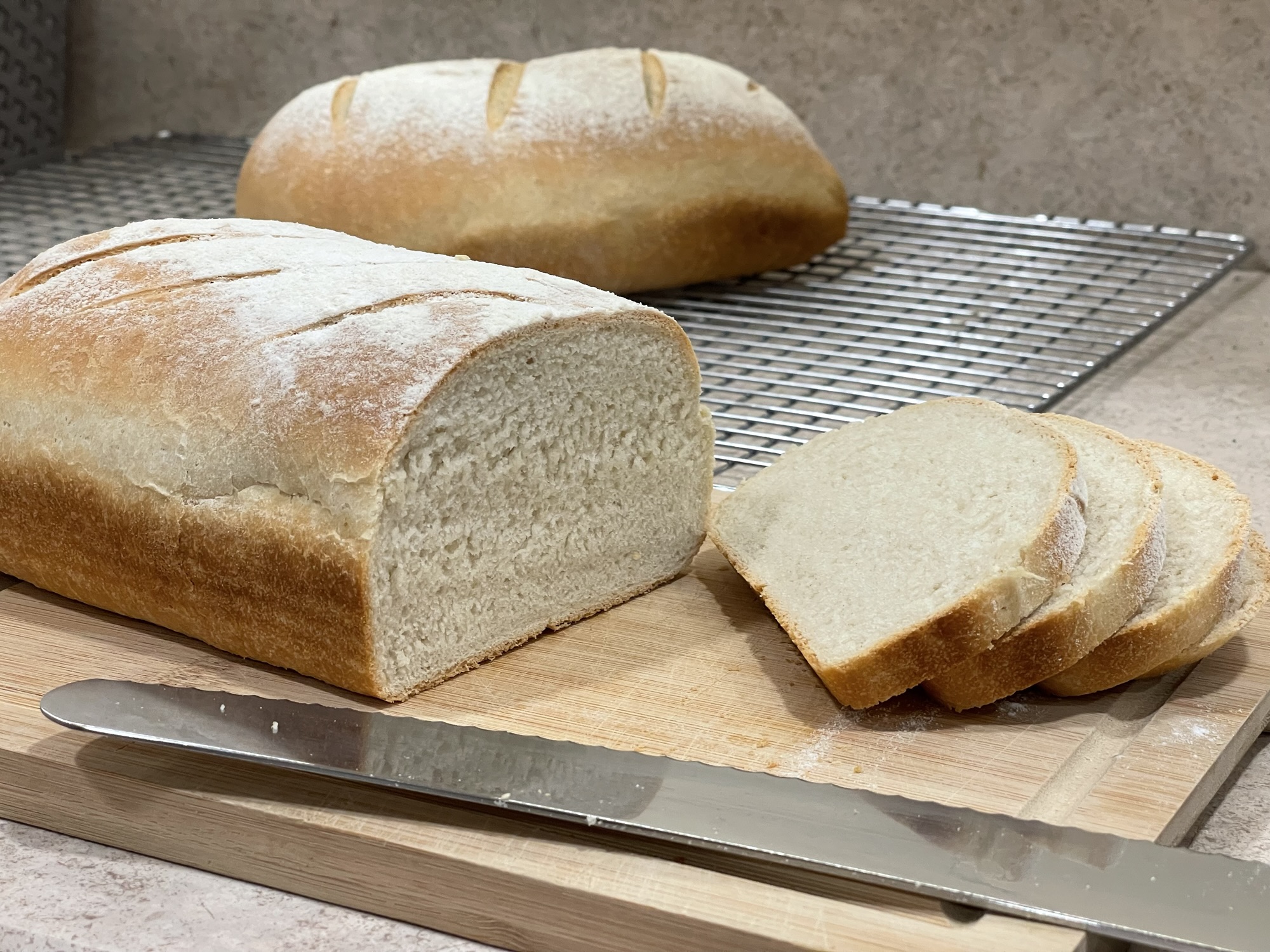 A load of bread on a wooden cutting board. It's partially sliced, with the slices lying to the side on the right. The serrated silver blade of a bread knife lies in front. Another uncut loaf is visible in the background.