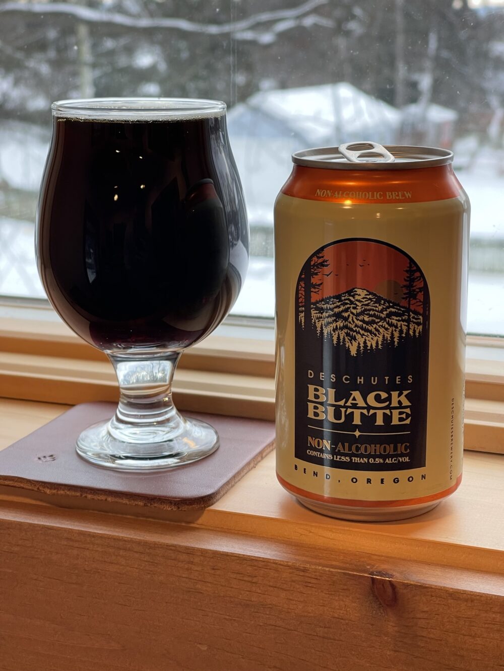 A tulip-shaped beer glass and a beer can sit on a windowsill. The glass is completely full of a deep brown, almost black liquid, with just a hint of light brown foam on top. The beer can next to it is beige and orange, with black accents. It shows a mountain covered with pine trees, and is labeled "Deschutes Black Butte Non-Alcoholic."