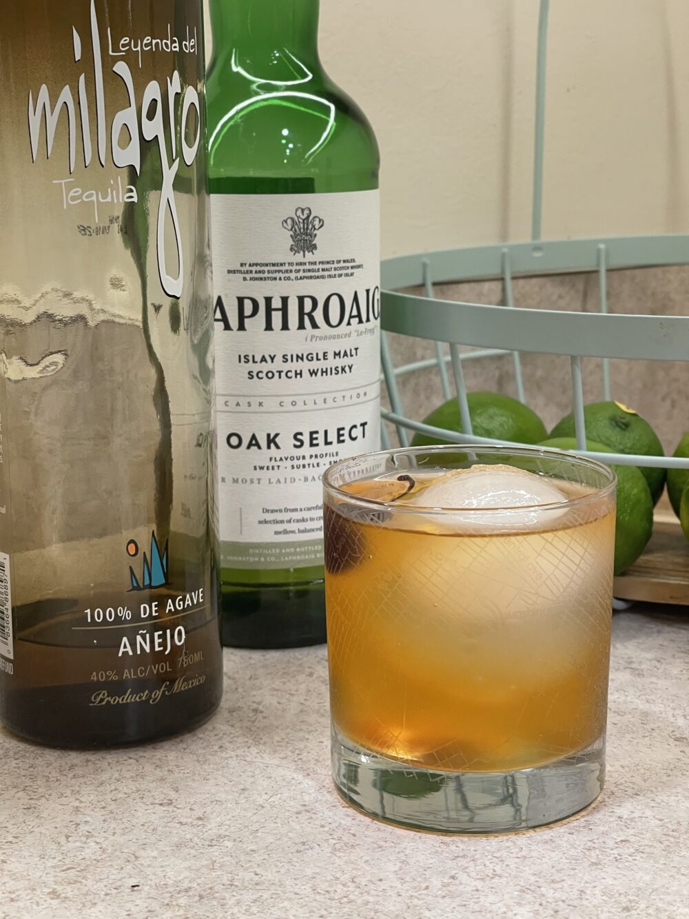 A cocktail in a rocks glass in front of some bottles and basket of fruit. The bottles are a green bottle of Laphroaig scotch and a deep olive green bottle of milagro tequila. The cocktail is a golden orange color. A ball of ice is in the glass, and a dark red cherry is visible towards the side.